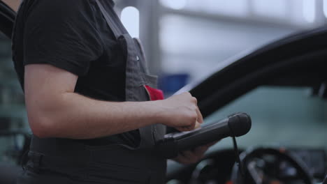 A-mechanic-in-a-close-up-car-service-holds-a-tablet-in-his-hands-and-presses-the-screen-near-the-car